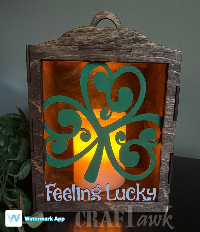 Pu&ograve; includere: Una lanterna di legno con un ritaglio a forma di trifoglio verde e il testo "Feeling Lucky" sul davanti. La lanterna &egrave; illuminata da una candela all'interno.