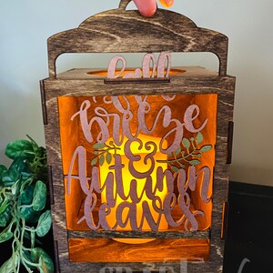 Pu&ograve; includere: Una lanterna di legno con una finitura colorata marrone e un pannello di vetro trasparente. La lanterna ha una maniglia sulla parte superiore e presenta un design ritagliato con il testo "Fall breeze & Autumn leaves".