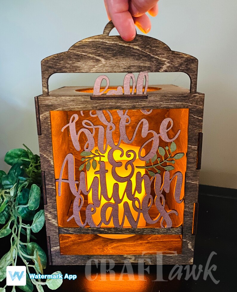 Pu&ograve; includere: Una lanterna di legno con una finitura colorata marrone e un pannello di vetro trasparente. La lanterna ha una maniglia sulla parte superiore e presenta un design ritagliato con il testo "Fall breeze & Autumn leaves".