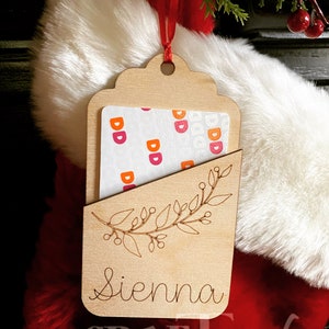 Puede incluir: Una etiqueta de regalo de madera con un recorte para una tarjeta de regalo. La etiqueta est&aacute; decorada con un dise&ntilde;o floral y el nombre "Sienna" est&aacute; grabado en la parte inferior. La etiqueta est&aacute; colgada de una cinta roja.