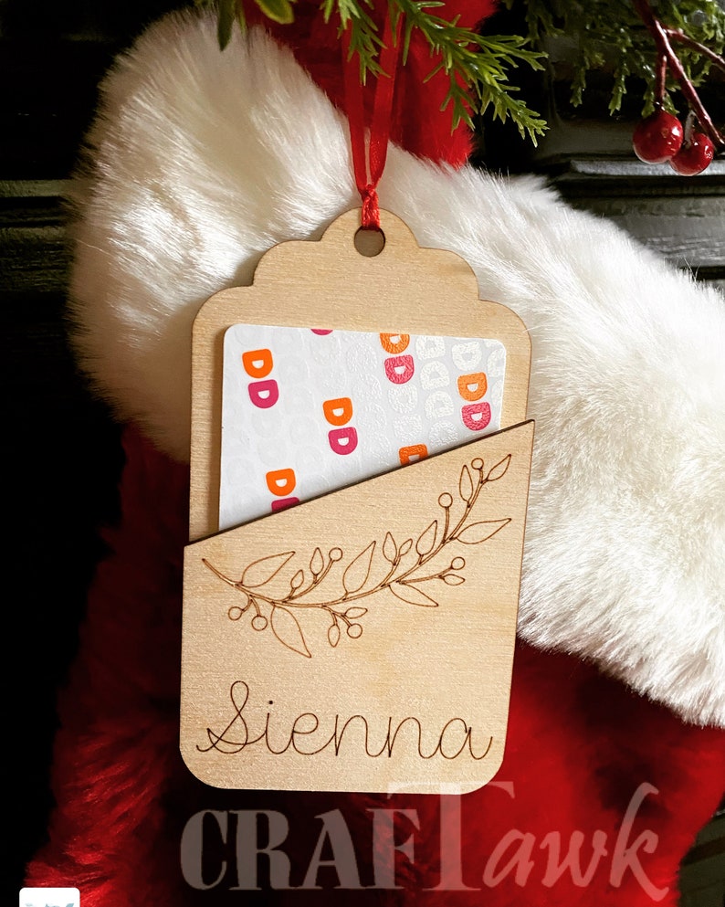 Puede incluir: Una etiqueta de regalo de madera con un recorte para una tarjeta de regalo. La etiqueta est&aacute; decorada con un dise&ntilde;o floral y el nombre "Sienna" est&aacute; grabado en la parte inferior. La etiqueta est&aacute; colgada de una cinta roja.