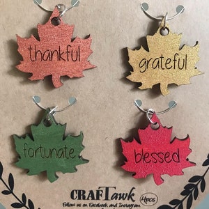 Puede incluir: Un conjunto de cuatro colgantes para vino con forma de hoja en colores oto&ntilde;ales: naranja, dorado, verde y rojo. Cada colgante tiene una palabra: "thankful", "grateful", "fortunate" y "blessed". El conjunto se presenta como un "wine charm set".