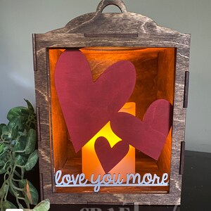 Pu&ograve; includere: Una lanterna di legno con una parte anteriore in vetro e una candela accesa all'interno. La lanterna contiene tre cuori di carta rossa all'interno, e il testo "love you more" &egrave; scritto sul vetro.