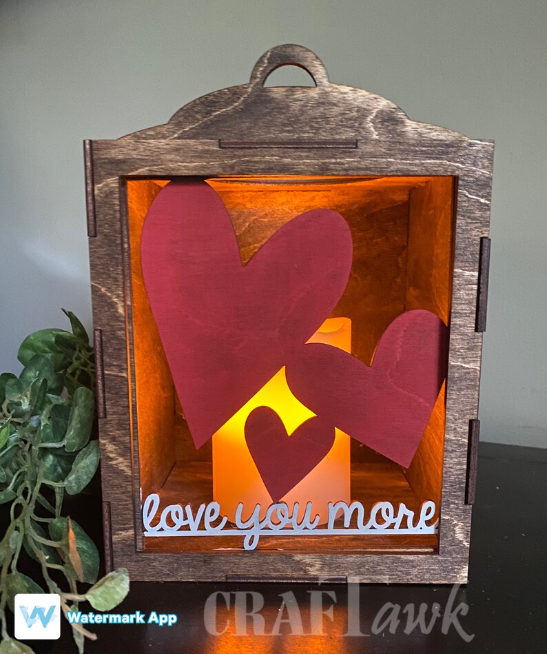 Pu&ograve; includere: Una lanterna di legno con una parte anteriore in vetro e una candela accesa all'interno. La lanterna contiene tre cuori di carta rossa all'interno, e il testo "love you more" &egrave; scritto sul vetro.