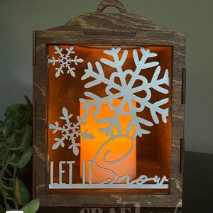 Pu&ograve; includere: Una lanterna di legno con una candela accesa all'interno. La lanterna ha un design a forma di fiocco di neve e le parole "Let it Snow".