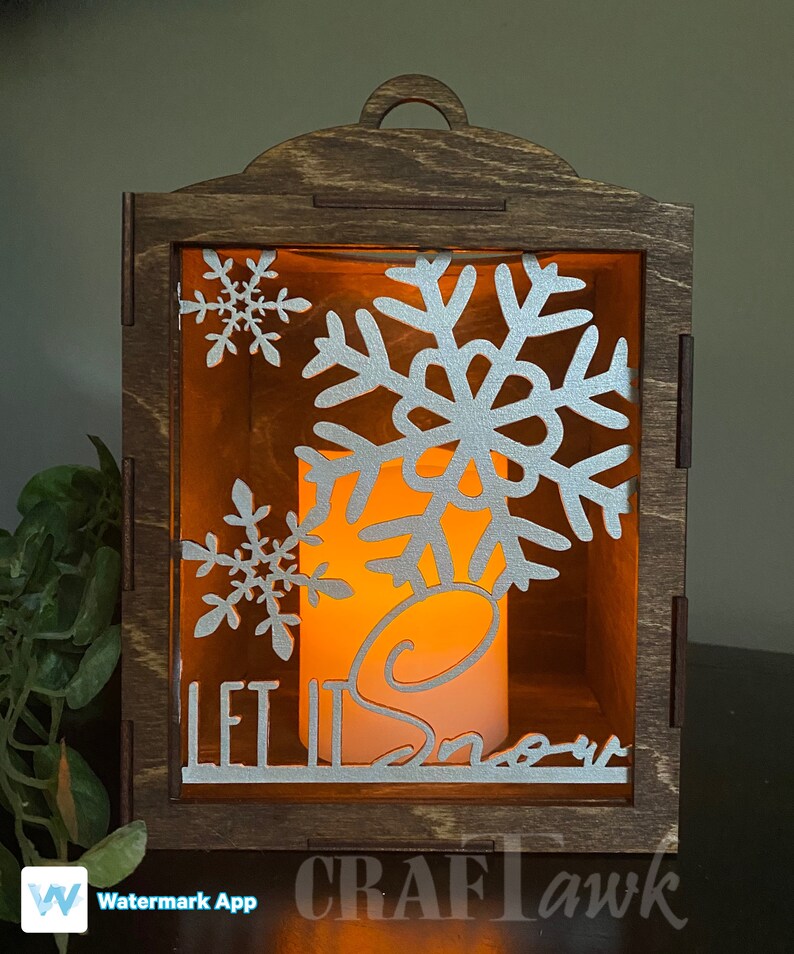 Pu&ograve; includere: Una lanterna di legno con una candela accesa all'interno. La lanterna ha un design a forma di fiocco di neve e le parole "Let it Snow".