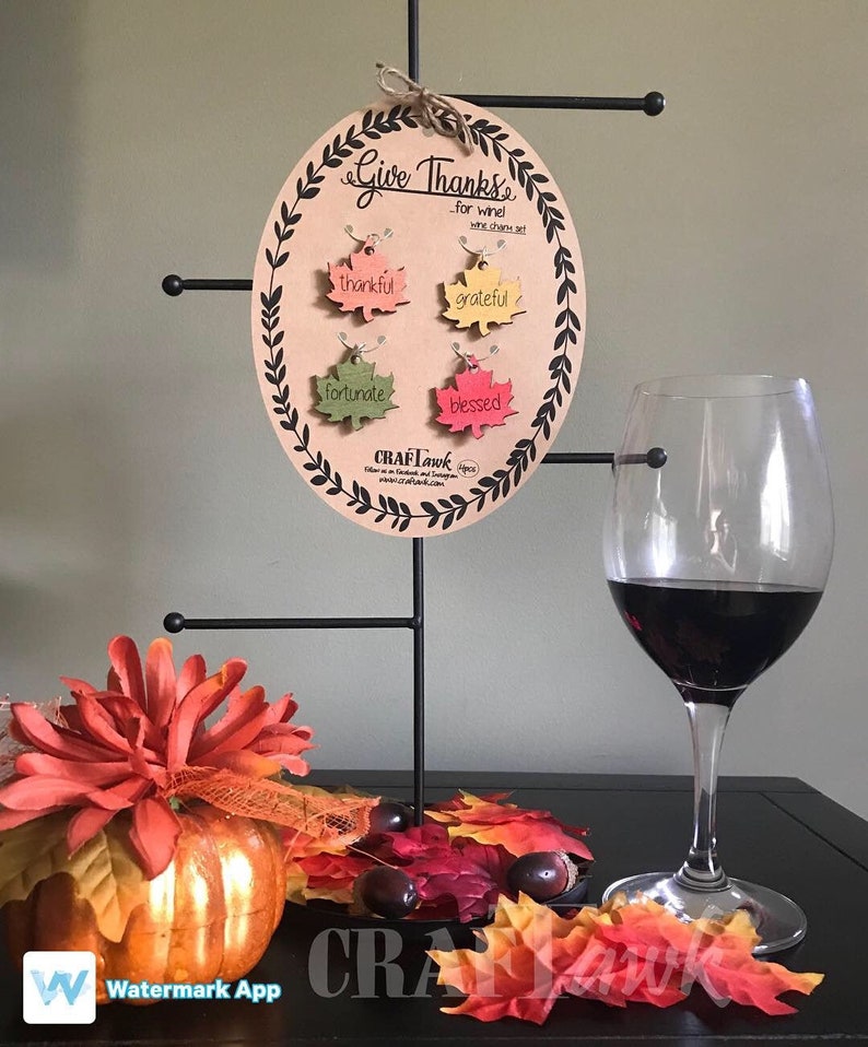 Puede incluir: Un juego de adornos para vino con las palabras "Give Thanks" y adornos en forma de hoja que dicen "thankful", "grateful", "fortunate" y "blessed". Una copa de vino tinto y decoraciones de oto&ntilde;o tambi&eacute;n est&aacute;n en la imagen.