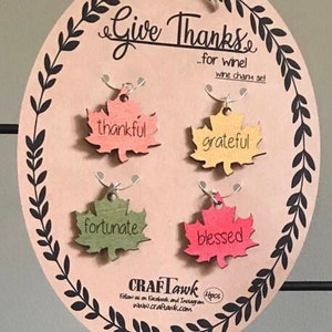 Puede incluir: Un juego de adornos para vino de madera con las palabras "Give Thanks" y cuatro adornos en forma de hoja. Cada adorno es de un color diferente y tiene una palabra: thankful, grateful, fortunate y blessed. El juego est&aacute; sobre un &oacute;valo marr&oacute;n claro con un borde de hojas negras.