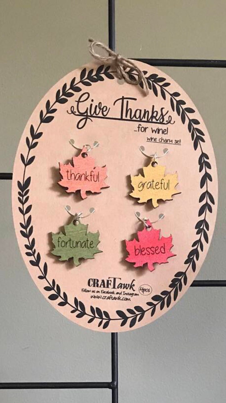 Puede incluir: Un juego de adornos para vino de madera con las palabras "Give Thanks" y cuatro adornos en forma de hoja. Cada adorno es de un color diferente y tiene una palabra: thankful, grateful, fortunate y blessed. El juego est&aacute; sobre un &oacute;valo marr&oacute;n claro con un borde de hojas negras.
