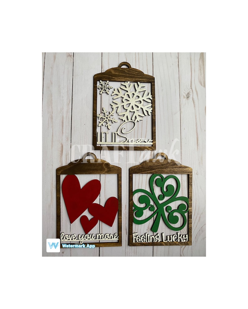 Pu&ograve; includere: Tre cartelli di legno con ritagli e testo. Il primo cartello ha un ritaglio a forma di fiocco di neve e il testo "Let it Snow". Il secondo cartello ha un ritaglio a forma di cuore e il testo "Love you more". Il terzo cartello ha un ritaglio a forma di trifoglio e il testo "Feeling Lucky".