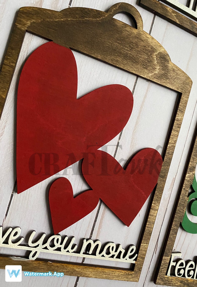 Pu&ograve; includere: Un cartello di legno con una cornice marrone e due cuori rossi. Il cartello dice "Ti amo di pi&ugrave;".