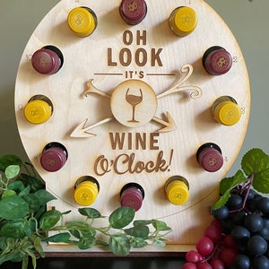 May include: A wooden wine o'clock clock with 12 slots for mini wine bottles. The clock face features the text "Oh Look It's Wine O'Clock!" with a wine glass icon. The clock is decorated with grapes and vines.