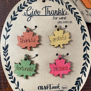 Puede incluir: Un juego de cuatro colgantes para vino con forma de hoja con las palabras "thankful", "grateful", "fortunate" y "blessed". Los colgantes est&aacute;n unidos a una tarjeta marr&oacute;n con el texto "Give Thanks...for wine!" y la marca "CRAFTawk".