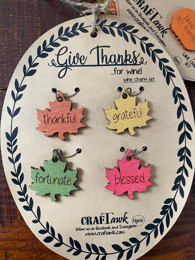 Puede incluir: Un juego de cuatro colgantes para vino con forma de hoja con las palabras "thankful", "grateful", "fortunate" y "blessed". Los colgantes est&aacute;n unidos a una tarjeta marr&oacute;n con el texto "Give Thanks...for wine!" y la marca "CRAFTawk".