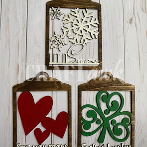 Pu&ograve; includere: Tre cartelli di legno con diversi disegni e detti. Il primo cartello &egrave; marrone con un fiocco di neve dorato e le parole "Let it Snow". Il secondo cartello &egrave; marrone con due cuori rossi e le parole "love you more". Il terzo cartello &egrave; marrone con un trifoglio verde e le parole "Feeling Lucky".