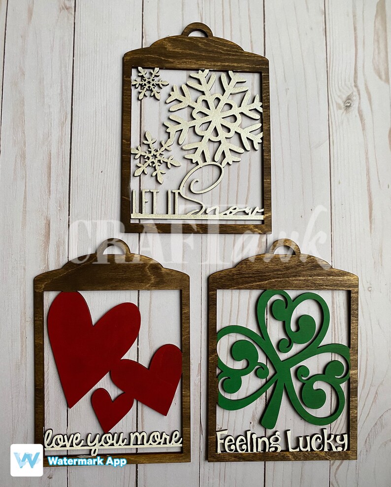 Pu&ograve; includere: Tre cartelli di legno con diversi disegni e detti. Il primo cartello &egrave; marrone con un fiocco di neve dorato e le parole "Let it Snow". Il secondo cartello &egrave; marrone con due cuori rossi e le parole "love you more". Il terzo cartello &egrave; marrone con un trifoglio verde e le parole "Feeling Lucky".