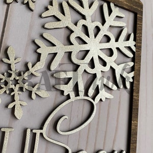 Pu&ograve; includere: Un cartello di legno con una finitura glitter argentata. Il cartello presenta un grande fiocco di neve e la parola "Neve" in un font corsivo.