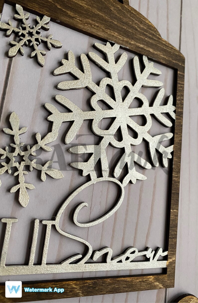 Pu&ograve; includere: Un cartello di legno con una finitura glitter argentata. Il cartello presenta un grande fiocco di neve e la parola "Neve" in un font corsivo.