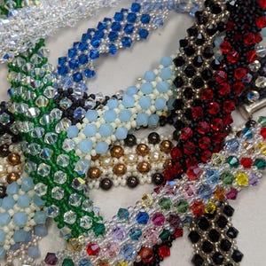 May include: A collection of beaded bracelets in various colors, including clear, blue, green, black, red, and multicolored. The bracelets feature different bead shapes and sizes, creating a textured and sparkling appearance. The bracelets are arranged closely together.