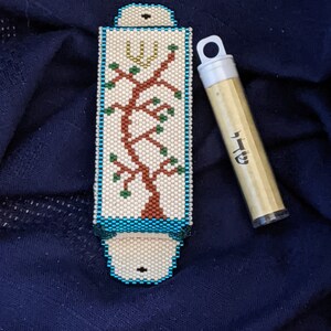 K&ouml;nnte beinhalten: Ein wei&szlig;er Mezuzah-Beh&auml;lter mit blauem Rand und einem Perlenmuster eines Baumes mit einem Davidstern oben. Der Beh&auml;lter befindet sich neben einem goldenen R&ouml;hrchen mit hebr&auml;ischer Schrift.