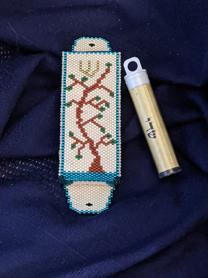 K&ouml;nnte beinhalten: Ein wei&szlig;er Mezuzah-Beh&auml;lter mit blauem Rand und einem Perlenmuster eines Baumes mit einem Davidstern oben. Der Beh&auml;lter befindet sich neben einem goldenen R&ouml;hrchen mit hebr&auml;ischer Schrift.