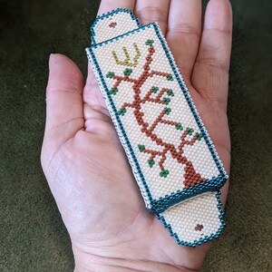 K&ouml;nnte beinhalten: Ein wei&szlig;er Perlen-Mezuzah-Beh&auml;lter mit einem gr&uuml;nen und braunen Baumdesign und einem goldenen hebr&auml;ischen Buchstaben.
