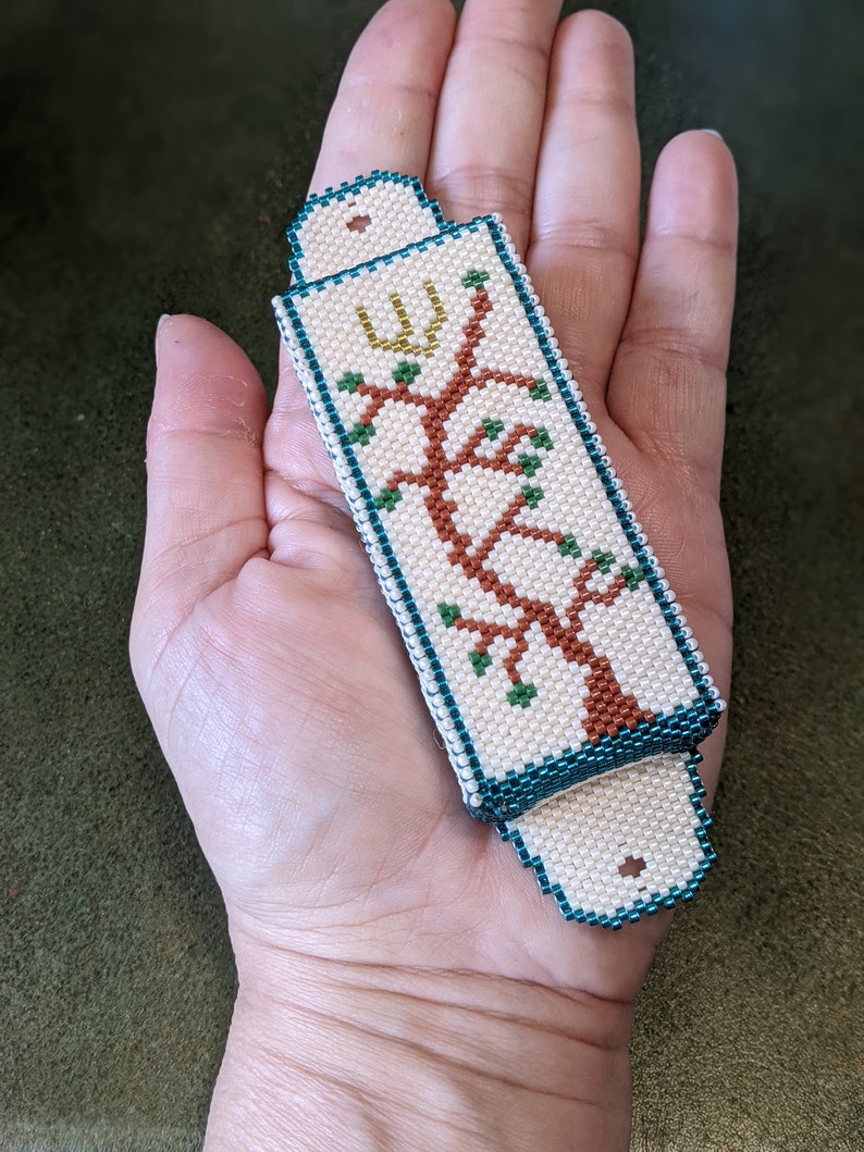 K&ouml;nnte beinhalten: Ein wei&szlig;er Perlen-Mezuzah-Beh&auml;lter mit einem gr&uuml;nen und braunen Baumdesign und einem goldenen hebr&auml;ischen Buchstaben.
