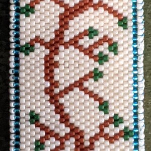 K&ouml;nnte beinhalten: Ein wei&szlig;er Perlen-Mezuzah-Beh&auml;lter mit blauem Rand. Der Beh&auml;lter zeigt ein Perlenmuster eines Baumes mit braunen &Auml;sten und gr&uuml;nen Bl&auml;ttern. Ein goldenes U befindet sich oben auf dem Baum.