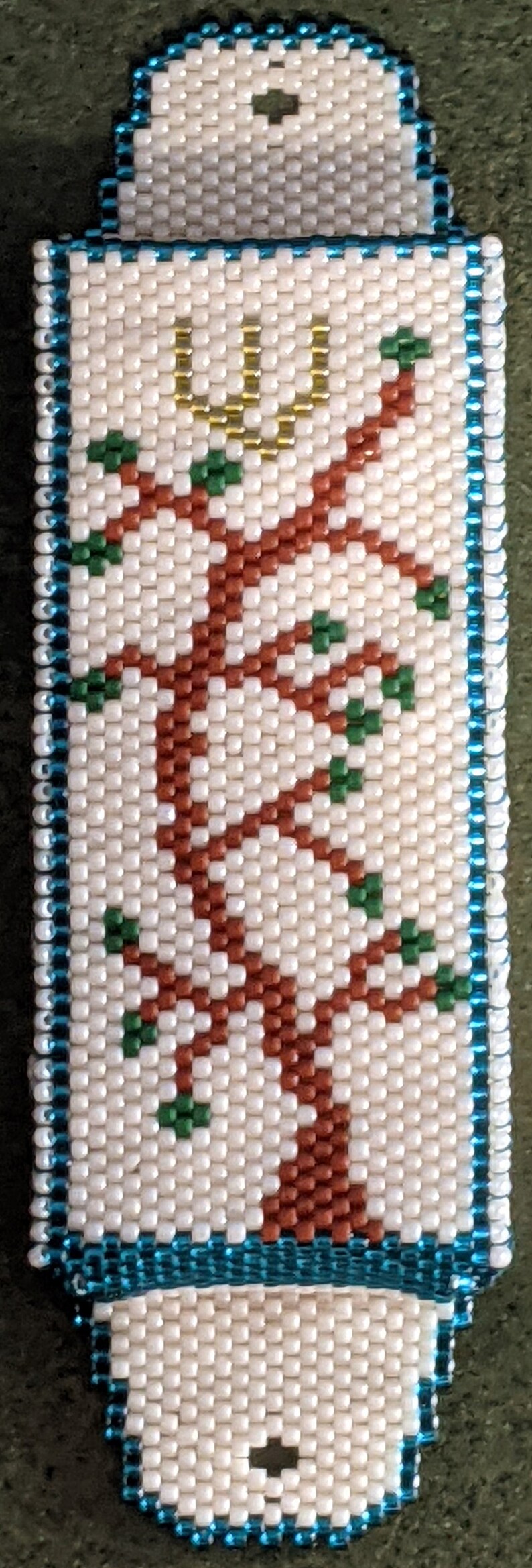 K&ouml;nnte beinhalten: Ein wei&szlig;er Perlen-Mezuzah-Beh&auml;lter mit blauem Rand. Der Beh&auml;lter zeigt ein Perlenmuster eines Baumes mit braunen &Auml;sten und gr&uuml;nen Bl&auml;ttern. Ein goldenes U befindet sich oben auf dem Baum.
