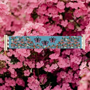 Könnte beinhalten: Ein Armband aus Perlen mit blauem Hintergrund und einem Blumenmuster. Drei rosa Schmetterlinge sind mittig auf dem Armband platziert. Das Armband hat goldene Metallverschlüsse.
