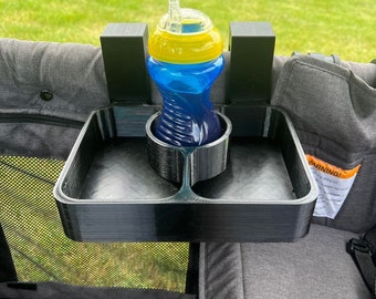 May include: A black plastic stroller tray with a built-in cup holder and a separate compartment. A blue and yellow sippy cup with a straw is in the cup holder. The tray is attached to a gray stroller.