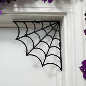 May include: Black metal spider web corner shelf bracket. The bracket is shaped like a spider web and is designed to be mounted in a corner.
