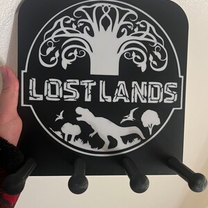 May include: Black wooden coat rack with four hooks. The rack features a white silhouette of a tree, a dinosaur, and the text "LOSTLANDS".