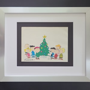Pode incluir: Uma ilustração emoldurada dos personagens dos Peanuts em pé em volta de uma árvore de Natal. Os personagens estão todos sorrindo e segurando presentes. A ilustração é assinada "SCHULZ" no canto inferior esquerdo.