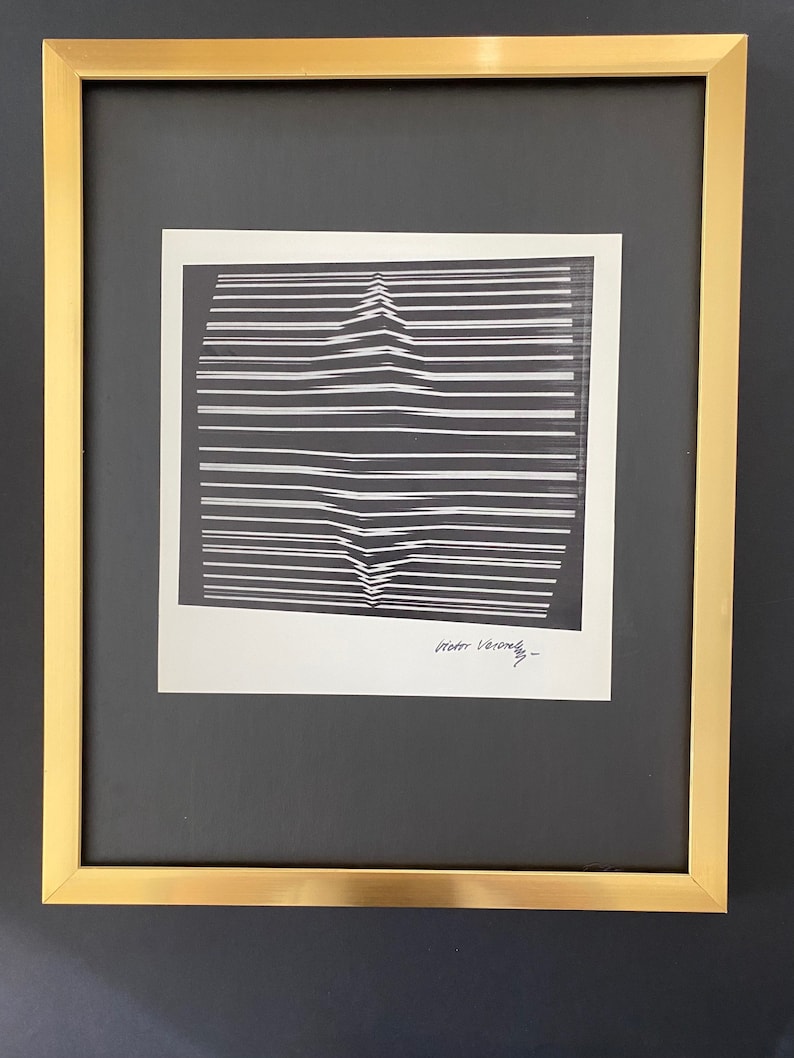 May include: A black and white abstract print framed in a gold frame. The print features a series of horizontal lines that create a visual illusion of depth and movement. The artist's signature, "Victor Vasarely", is visible in the bottom right corner of the print.