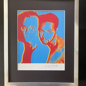 Puede incluir: Una impresi&oacute;n enmarcada de un retrato pop art de dos hombres, uno con gafas, en colores rojo, amarillo y azul. La impresi&oacute;n est&aacute; firmada "Andy Warhol".