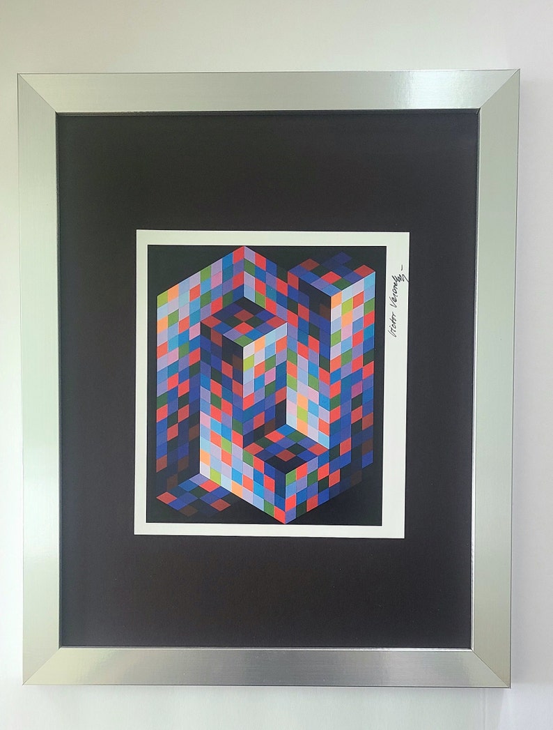 May include: A framed print of a colorful geometric abstract art piece. The artwork is a multi-colored cube with a black background. The artist's signature, "Victor Vasarely", is in the bottom right corner of the print.