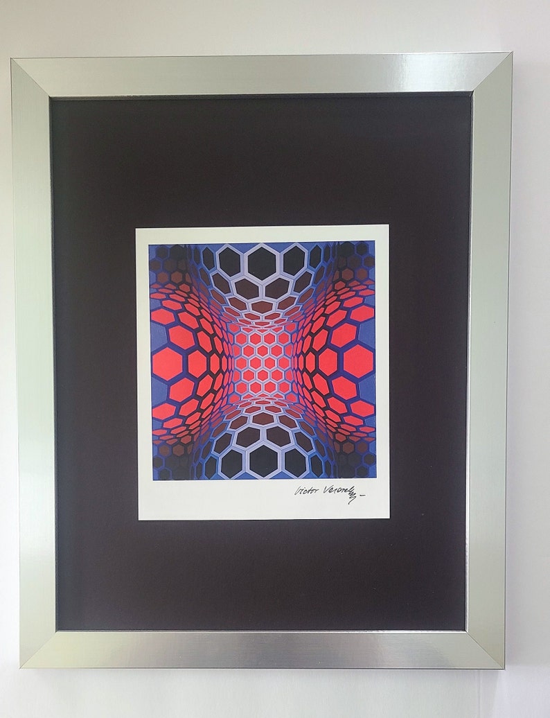 Op de afbeelding: Een ingelijste print van een geometrische optische illusie met een rood, blauw en zwart honingraatpatroon. De print is gesigneerd "Victor Vasarely".