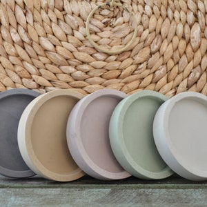 May include: Five round concrete coasters in different colors: gray, beige, light purple, light green, and white. The coasters are arranged in a row on a wooden surface.