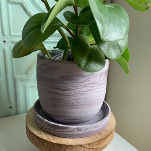 May include: A small potted plant with green leaves in a purple marbled ceramic pot. The pot is sitting on a wooden stand with three legs.