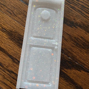 May include: A white door-shaped ornament with iridescent glitter embedded in the resin. The ornament has a small round knob and a rectangular window.