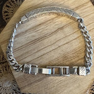 K&ouml;nnte beinhalten: Ein silbernes Armband mit einem Panzerketten-Design und einem dekorativen rechteckigen Mittelst&uuml;ck. Das Mittelst&uuml;ck weist ein eingraviertes Muster auf. Das Armband wird mit einem Verschluss gesichert. Das Armband wird auf einer Holzoberfl&auml;che pr&auml;sentiert.