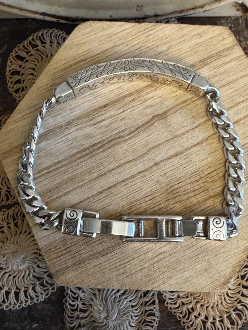 K&ouml;nnte beinhalten: Ein silbernes Armband mit einem Panzerketten-Design und einem dekorativen rechteckigen Mittelst&uuml;ck. Das Mittelst&uuml;ck weist ein eingraviertes Muster auf. Das Armband wird mit einem Verschluss gesichert. Das Armband wird auf einer Holzoberfl&auml;che pr&auml;sentiert.