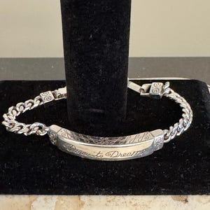 K&ouml;nnte beinhalten: Ein silbernes Armband mit einem Panzerketten-Design. Der Mittelsteg ist mit den Worten "Dare to Dream" in einer Schreibschrift graviert. Das Armband wird auf einer schwarzen Oberfl&auml;che pr&auml;sentiert.