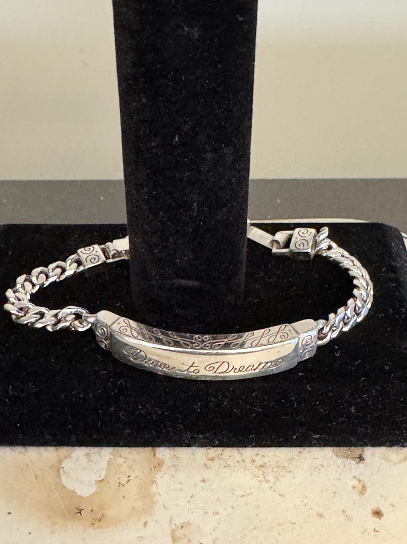 K&ouml;nnte beinhalten: Ein silbernes Armband mit einem Panzerketten-Design. Der Mittelsteg ist mit den Worten "Dare to Dream" in einer Schreibschrift graviert. Das Armband wird auf einer schwarzen Oberfl&auml;che pr&auml;sentiert.