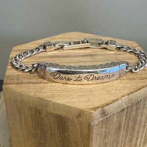 K&ouml;nnte beinhalten: Silberfarbenes Armband mit Panzerketten-Design. Das Mittelst&uuml;ck tr&auml;gt den eingravierten Schriftzug "Dare to Dream" in Schreibschrift. Das Armband wird auf einem Holzblock pr&auml;sentiert, um sein Design hervorzuheben.