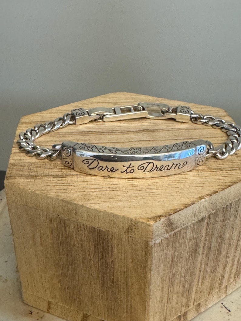 K&ouml;nnte beinhalten: Silberfarbenes Armband mit Panzerketten-Design. Das Mittelst&uuml;ck tr&auml;gt den eingravierten Schriftzug "Dare to Dream" in Schreibschrift. Das Armband wird auf einem Holzblock pr&auml;sentiert, um sein Design hervorzuheben.