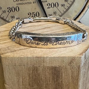 K&ouml;nnte beinhalten: Silbernes Armband mit einem rechteckigen Mittelst&uuml;ck, das mit den Worten "Dare to Dream" in Schreibschrift graviert ist. Das Armband hat ein Kettenglied-Design und wird auf einem Holzblock pr&auml;sentiert.