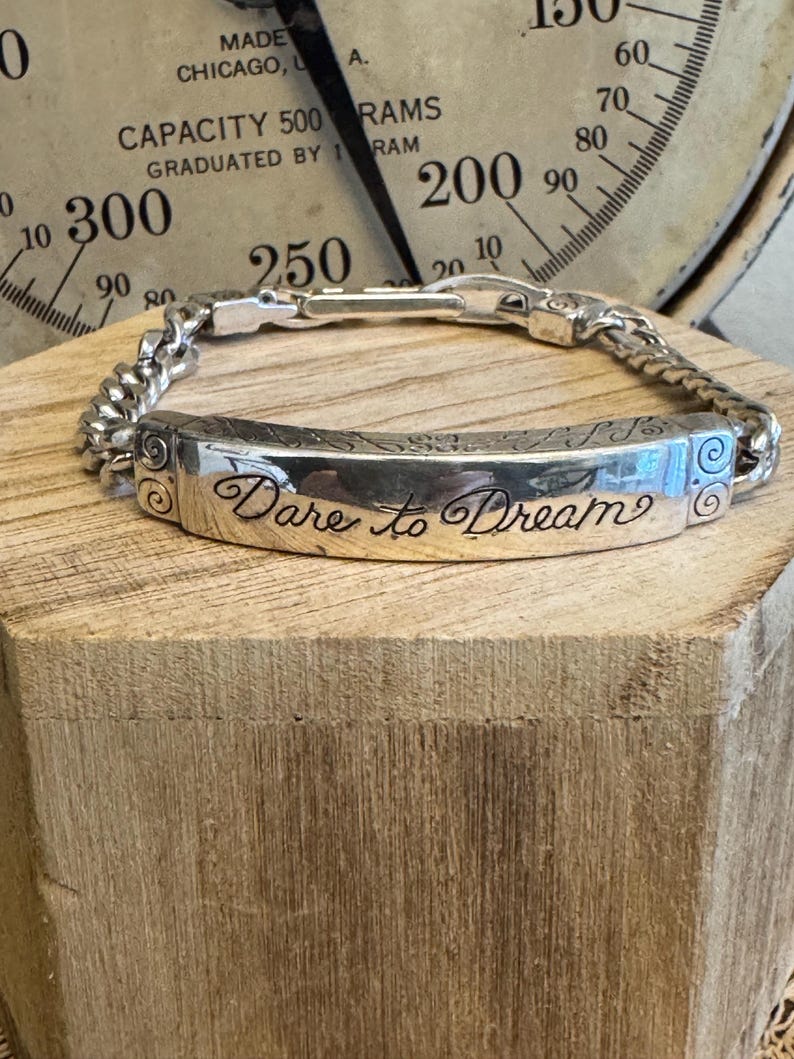 K&ouml;nnte beinhalten: Silbernes Armband mit einem rechteckigen Mittelst&uuml;ck, das mit den Worten "Dare to Dream" in Schreibschrift graviert ist. Das Armband hat ein Kettenglied-Design und wird auf einem Holzblock pr&auml;sentiert.
