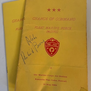 May include: Yellow paper document with the title "Change of Command" and the text "Fleet Marine Force Pacific" in red. The document is signed "Aloha Phil and Berry" and has a red seal with the text "FMF-PAC" and three stars. The document also includes the text "at the Marine Corps Air Station Kaneohe Bay, Oahu, Hawaii 31 May 1968."
